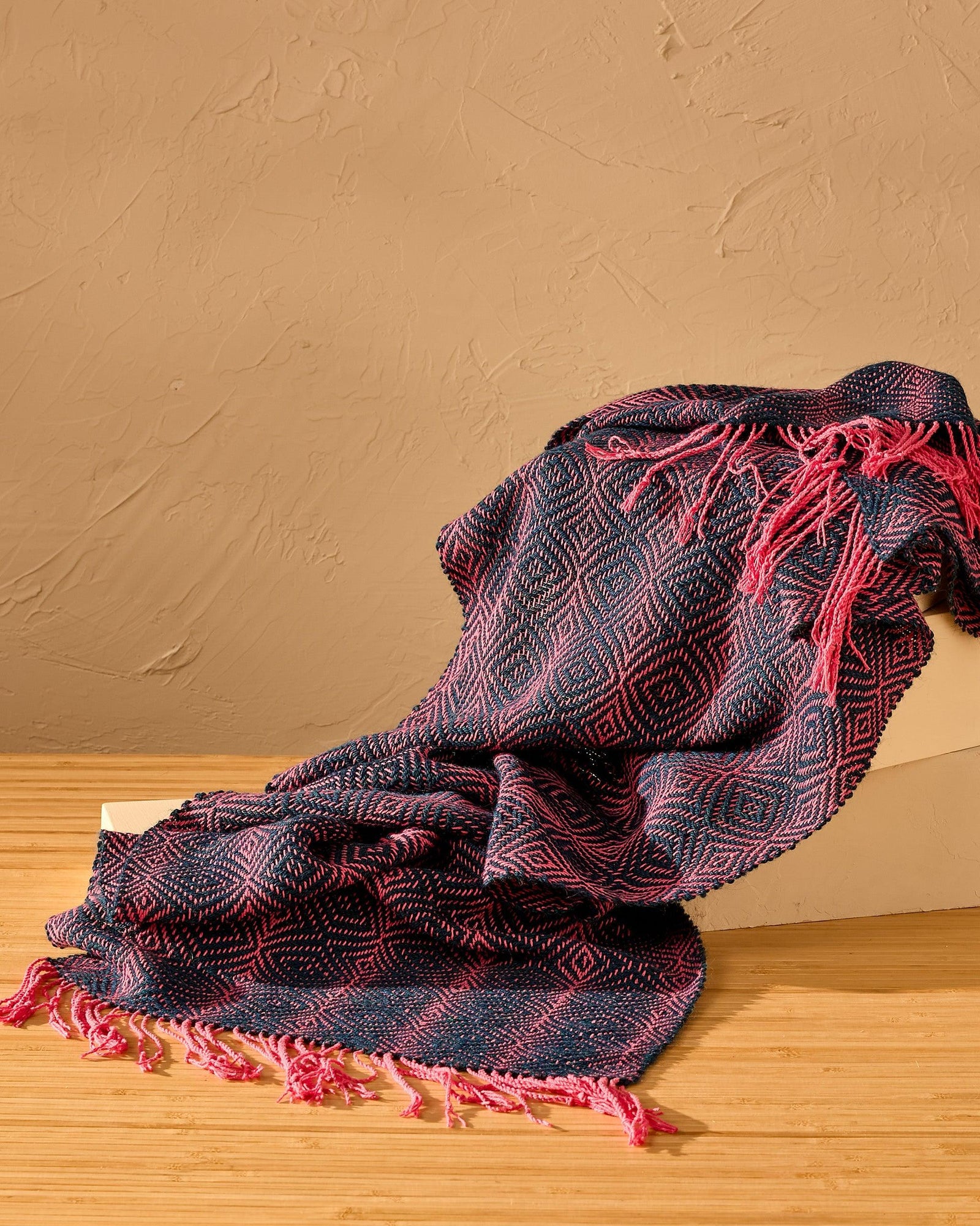 Woven scarf draped over a wooden surface against a beige wall.