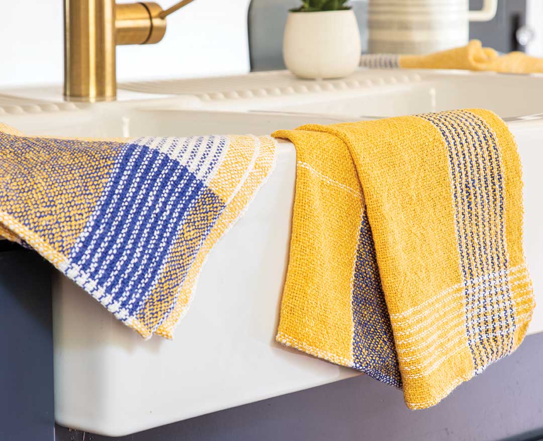 Amalfi Coast Towels in blue and yellow woven patterns displayed over a white kitchen sink with gold faucet.