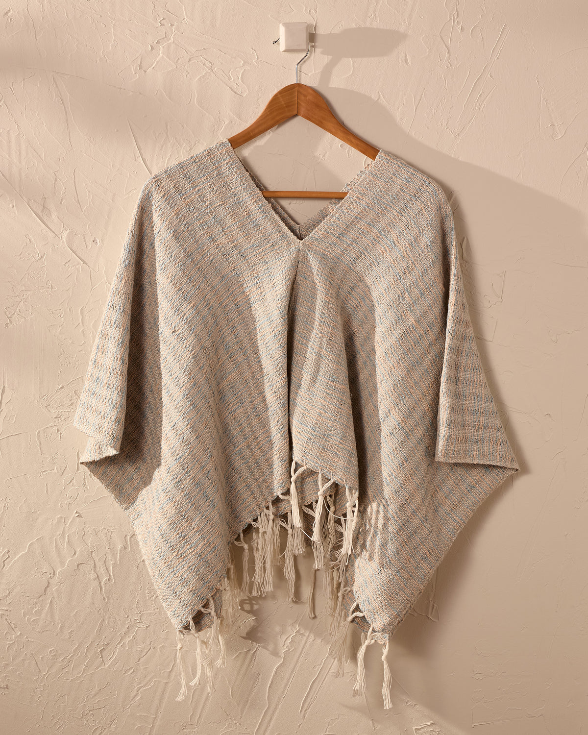 handwoven poncho with fringe on a wooden hanger against a beige wall